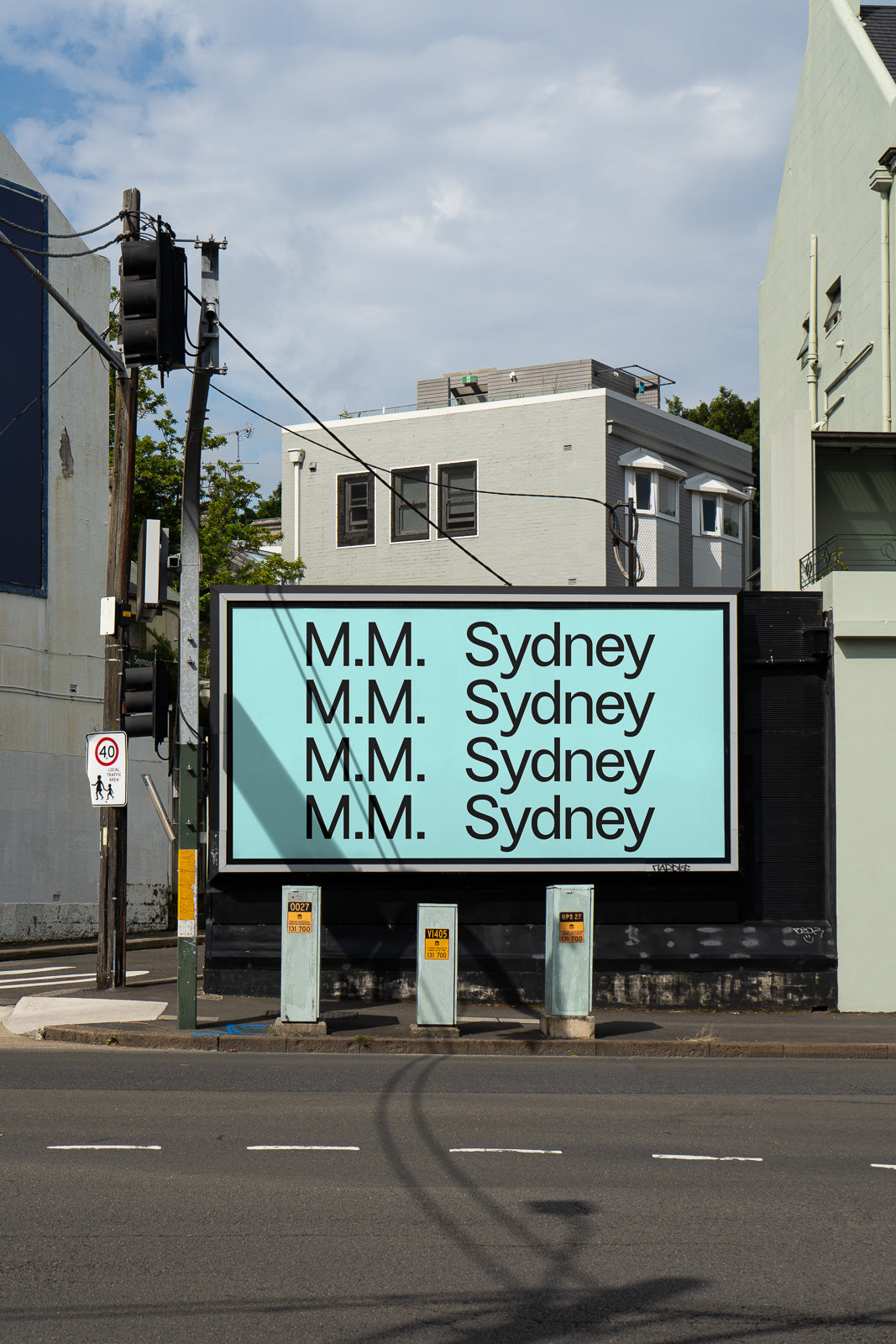 PSD mockup of a large outdoor billboard displaying repeated text "M.M. Sydney" in bold black letters on a light blue background, set against an urban street scene.