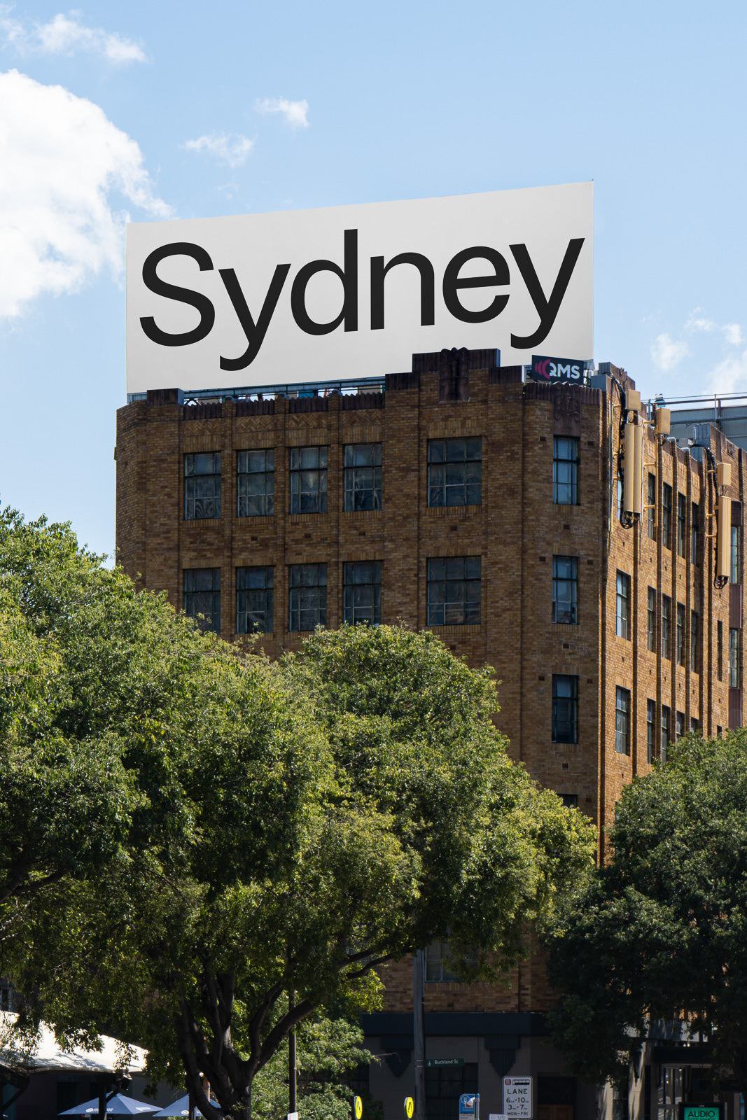 PSD mockup of a large billboard with "Sydney" written on it, placed atop a brick building with trees in the foreground and a clear sky above.