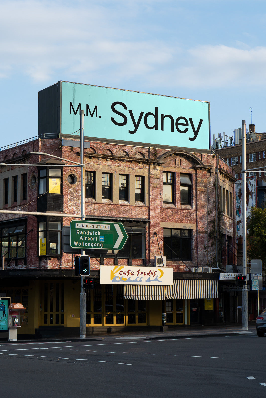 PSD mockup of a large rectangular billboard on a vintage brick building, displaying "M.M. Sydney" in bold text against a pastel blue background.