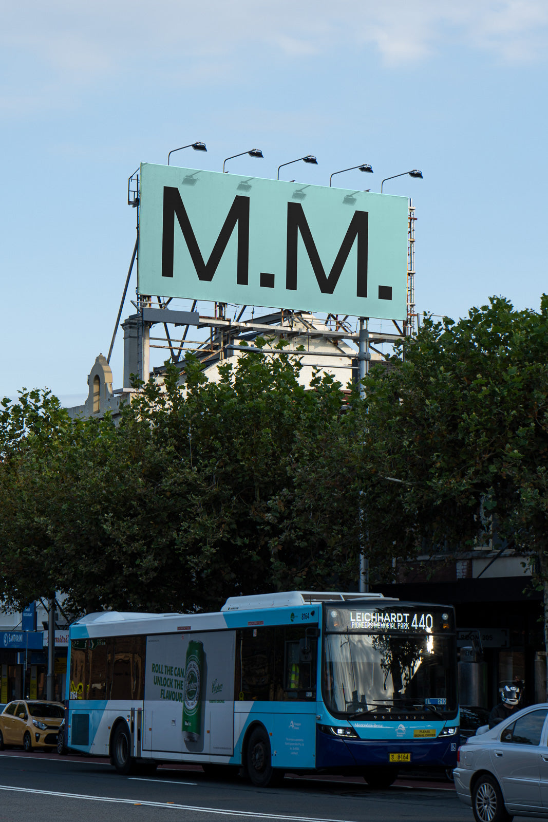PSD mockup of a large billboard displaying "M.M." in bold black letters against a light blue sky, with a public bus passing beneath it.