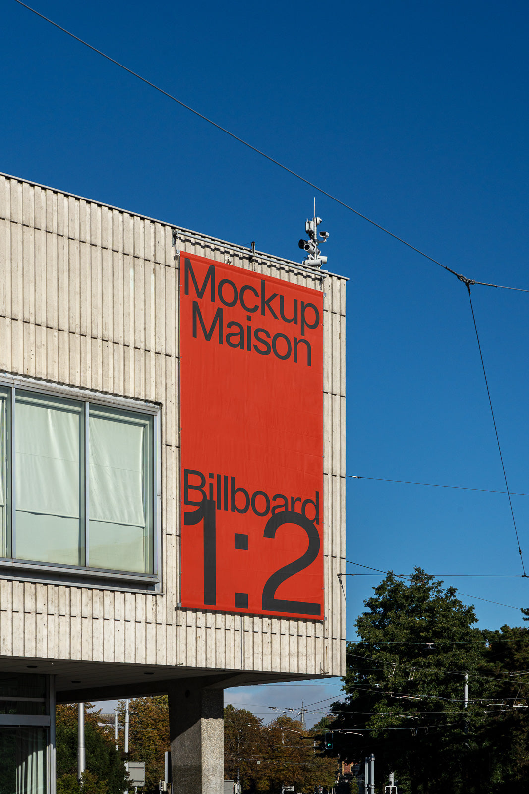 PSD mockup of a large red billboard reading "Mockup Maison Billboard 1:2" on the side of a concrete building with a clear blue sky.