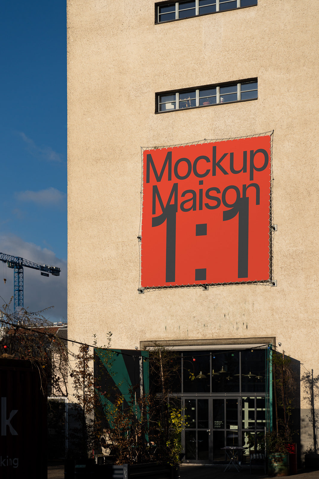 PSD mockup of a large red banner with "Mockup Maison 1:1" text on the wall of a building, with a crane and trees in the background.