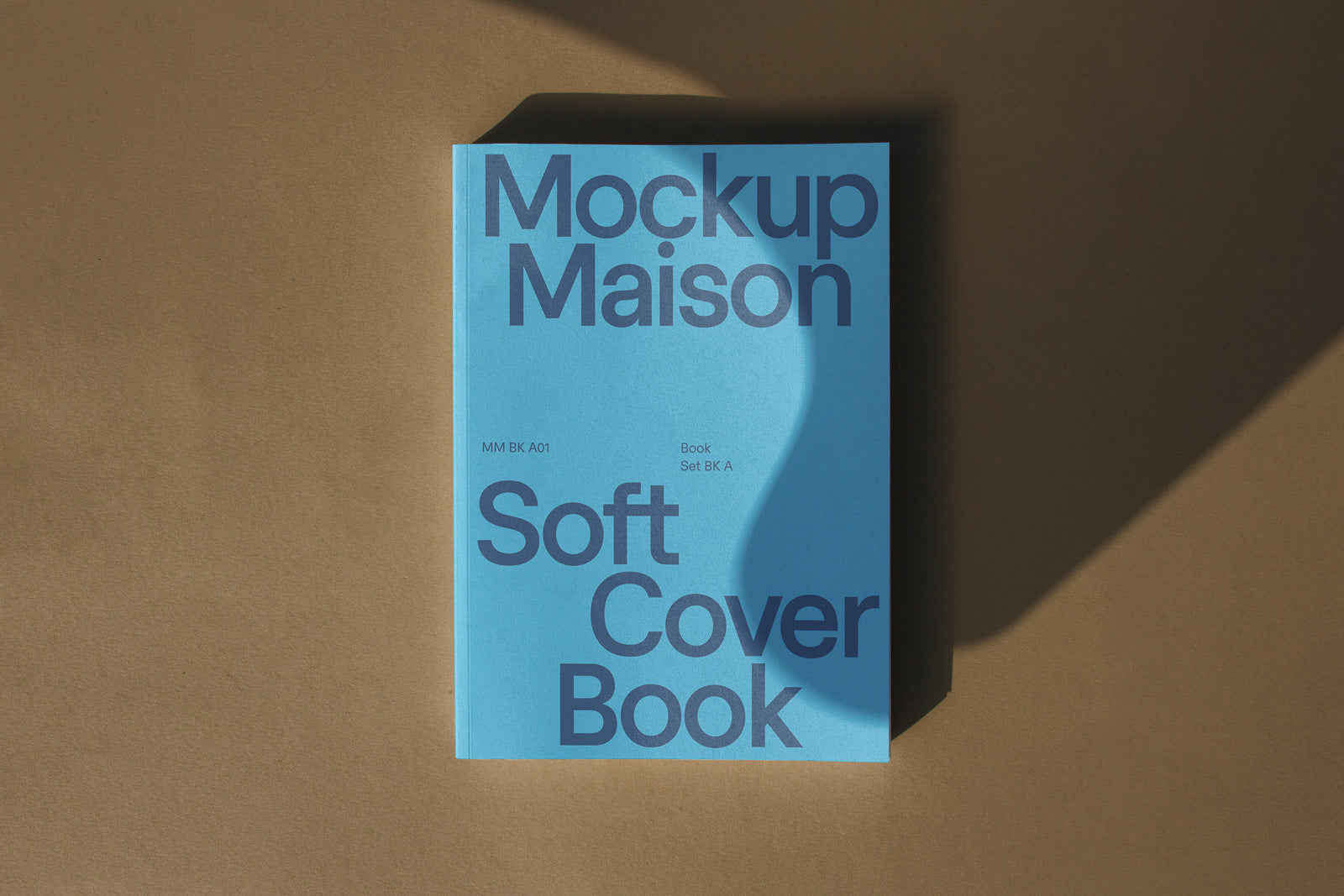 PSD mockup of a soft cover book with "Mockup Maison" text in bold on a blue cover, casting a shadow on a brown surface.