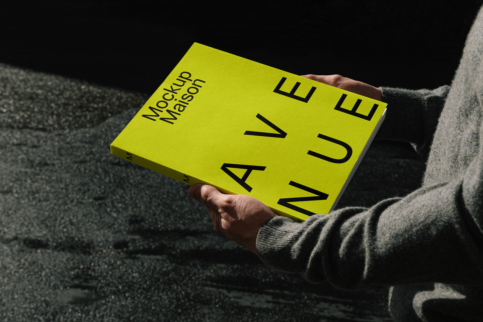 PSD mockup of a person holding a bright yellow book with "Mockup Maison" and "AVENUE" in bold black text, casting shadows on a textured surface.