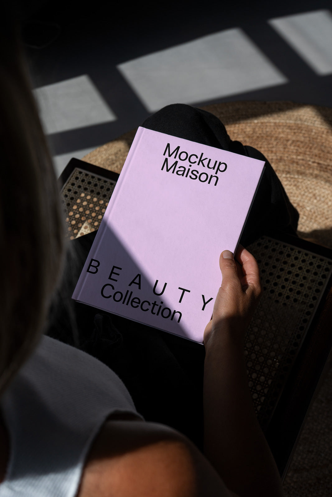 PSD mockup of a person holding a purple book titled "Mockup Maison Beauty Collection," with sunlight casting shadows on a wicker chair.