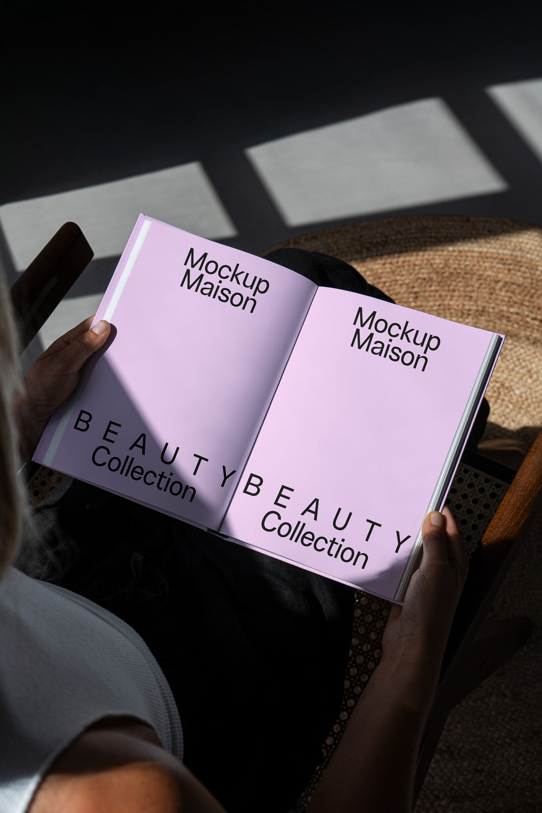 PSD mockup of a person holding a book with the title "Mockup Maison Beauty Collection" on pink pages, in a shadowed setting.