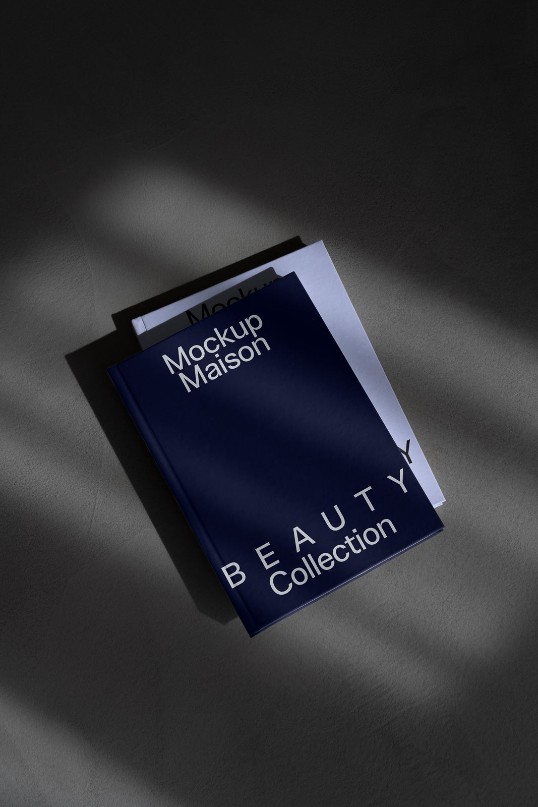 PSD mockup of a minimalistic dark blue and white book cover titled "Mockup Maison Beauty Collection" on a textured surface with shadows.