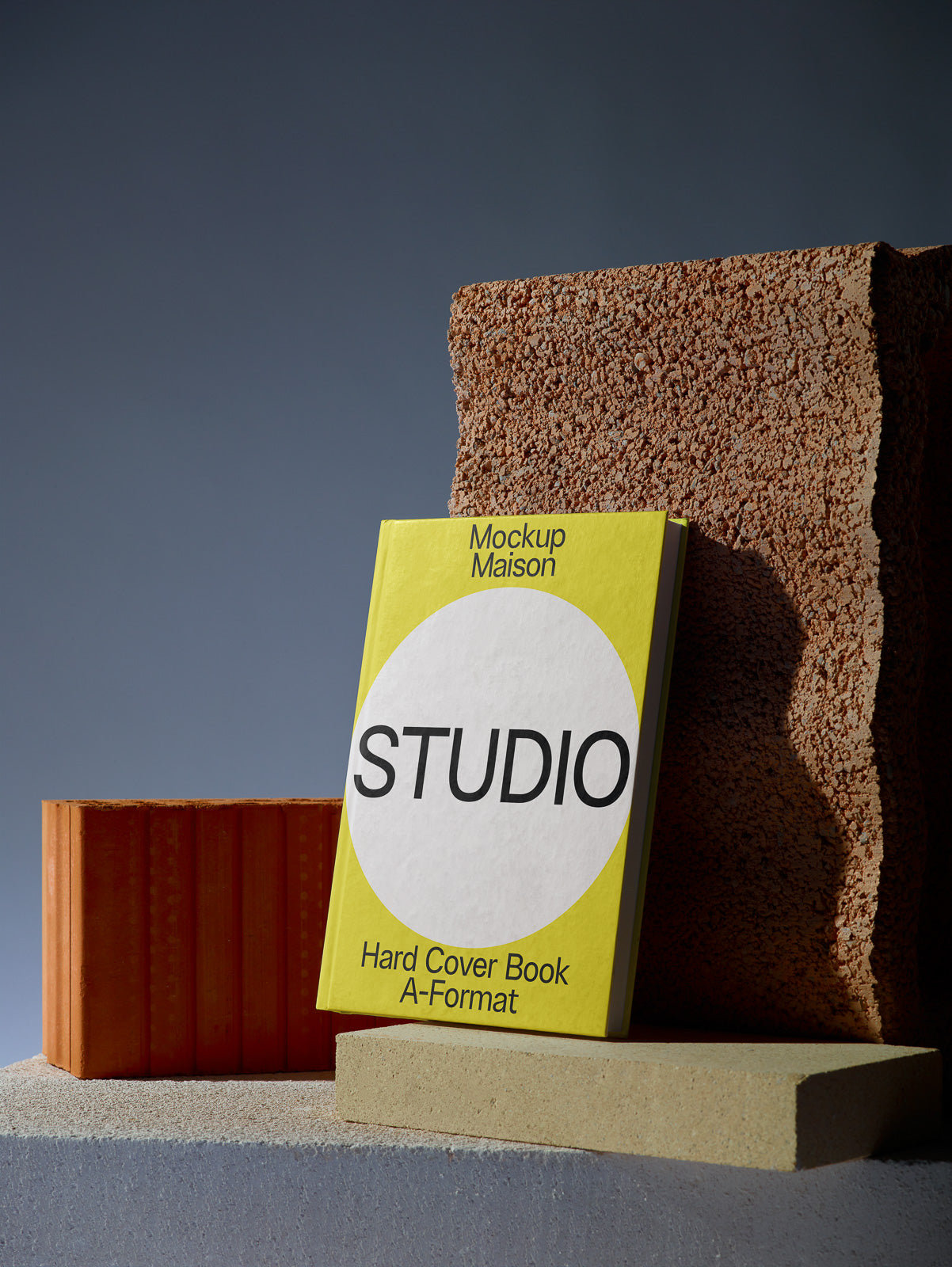 PSD mockup of a yellow hardcover book titled "STUDIO" with textured blocks in the background, set on a concrete surface.