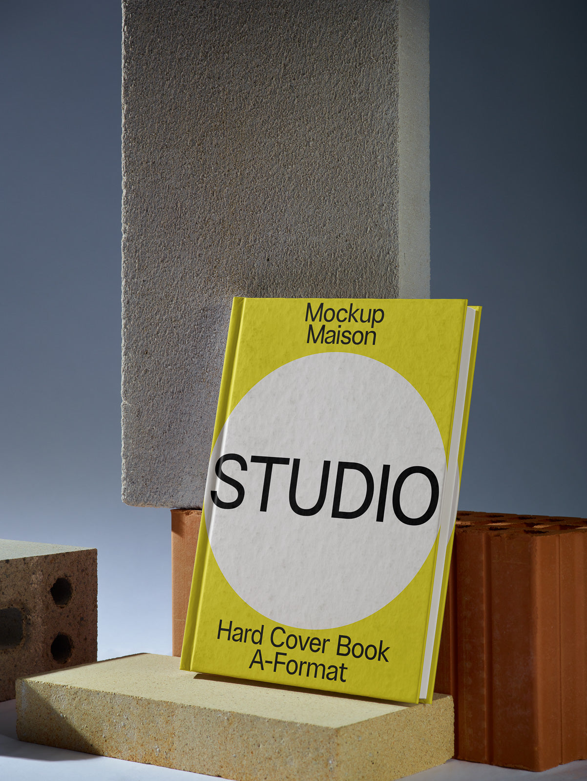 PSD mockup of a yellow hardcover book titled "Mockup Maison Studio," set against a backdrop of various concrete blocks.