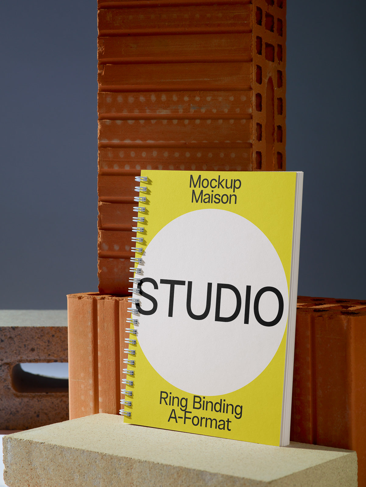 PSD mockup of a spiral-bound notebook with a yellow and white cover labeled "Studio" against a backdrop of stacked bricks.