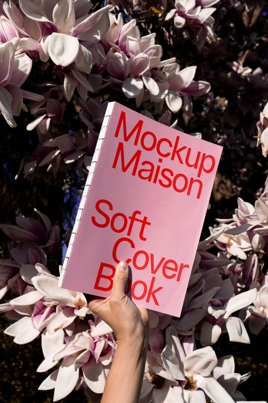 PSD mockup of a hand holding a pink soft cover book with red text, set against a backdrop of blooming magnolia flowers.
