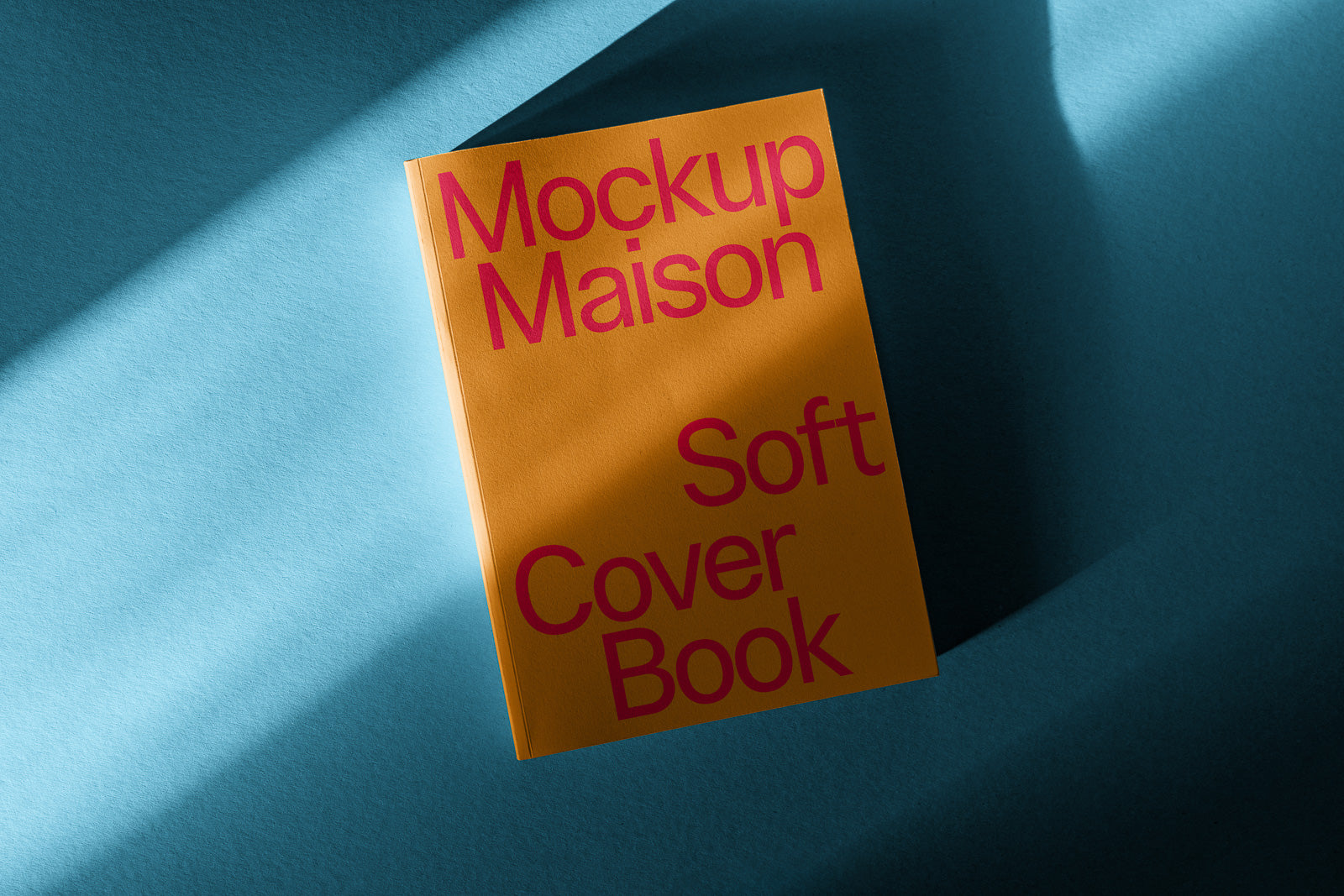 PSD mockup of a soft cover book titled "Mockup Maison," with bold pink text on an orange cover, resting on a textured blue surface with light shadows.