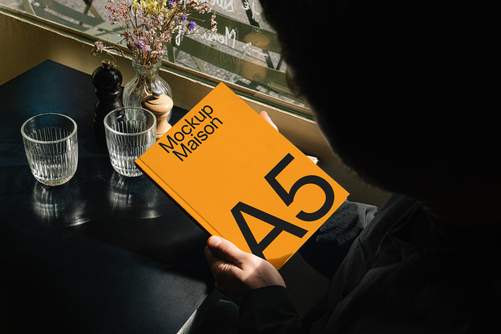 PSD mockup of person holding an A5-sized orange booklet titled "Mockup Maison," seated at a table with glasses and a vase of flowers nearby.