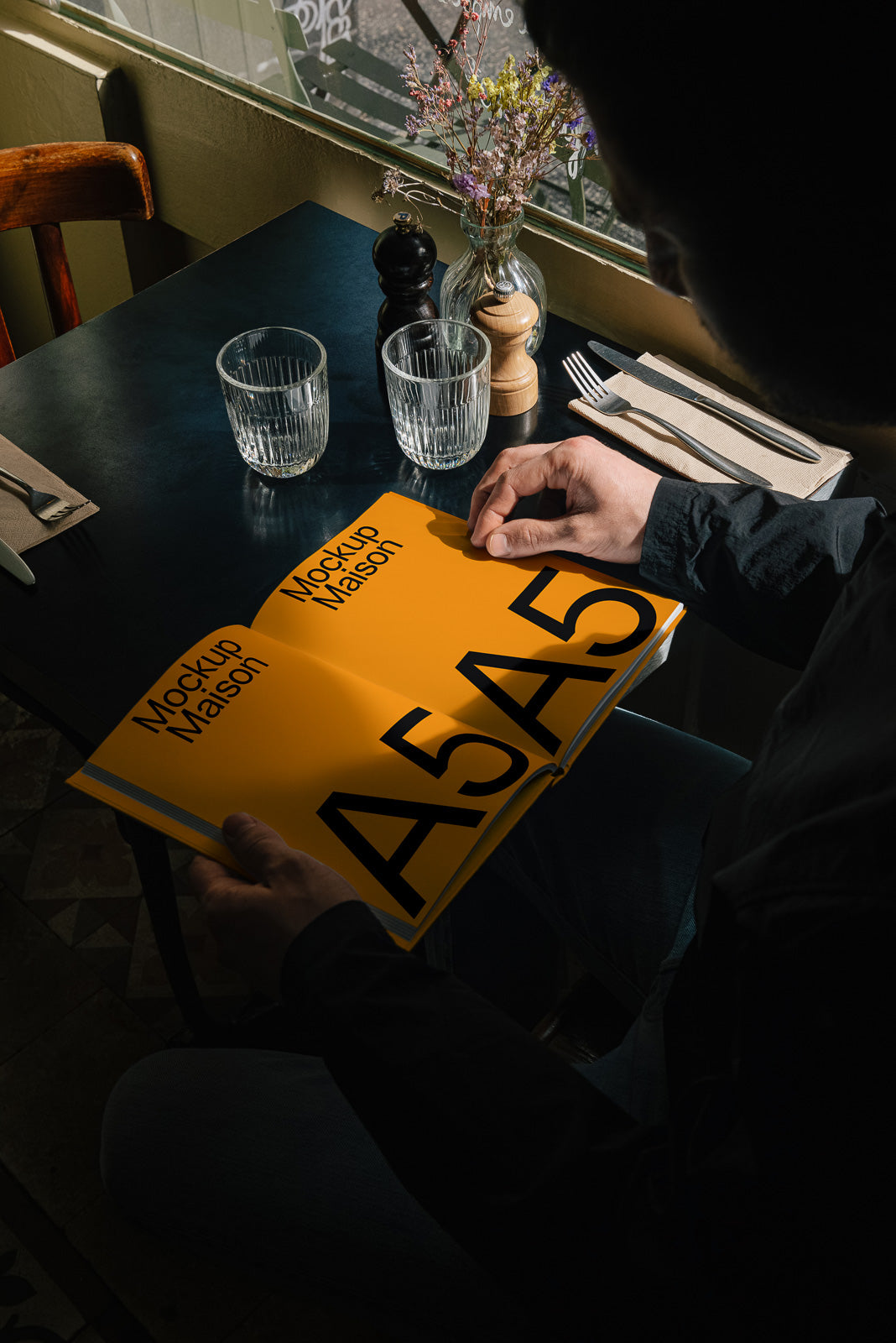 PSD mockup of a person holding an open A5 booklet with a bold yellow cover and black lettering in a dimly lit cafe setting.