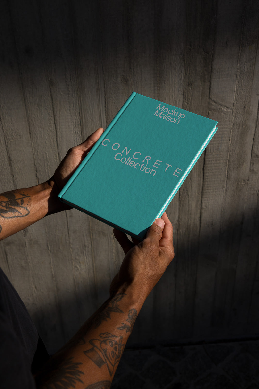 PSD mockup of a person holding a turquoise book titled "Concrete Collection" against a textured concrete wall background, casting soft shadows.