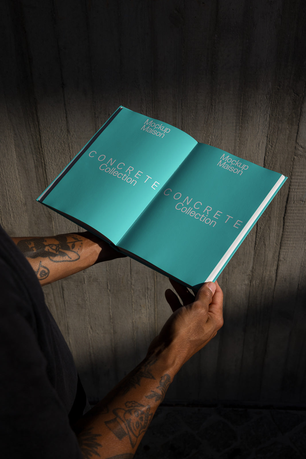 PSD mockup of a person holding an open book with the text "Concrete Collection" on the cyan pages, showcasing tattooed arms against a textured background.