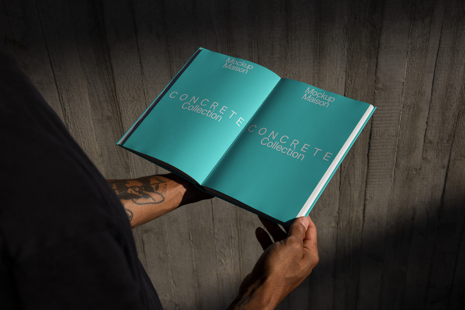 PSD mockup of a person holding an open book with a teal cover titled "Concrete Collection" and "Mockup Maison," against a dark wooden background.