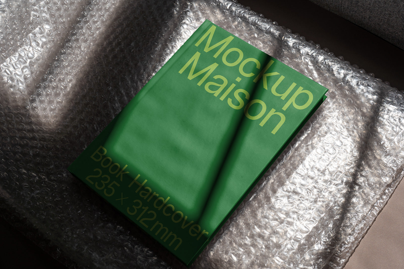 PSD mockup of a green hardcover book with title "Mockup Maison" on bubble wrap, showcasing dimensions 235 x 312mm in sunlight.