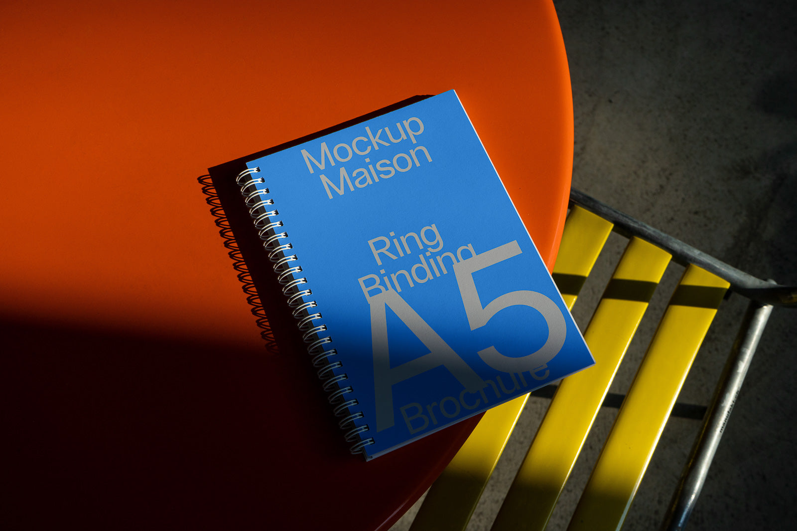 PSD mockup of an A5 ring-bound brochure with a blue cover on an orange surface, alongside a yellow metal chair.