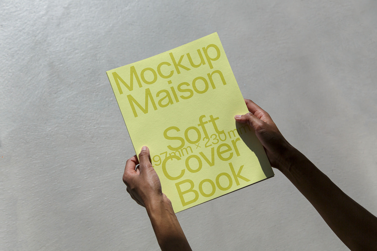 PSD mockup of hands holding a softcover book with a light green cover and bold text: "Mockup Maison Soft Cover Book 197mm x 230mm."