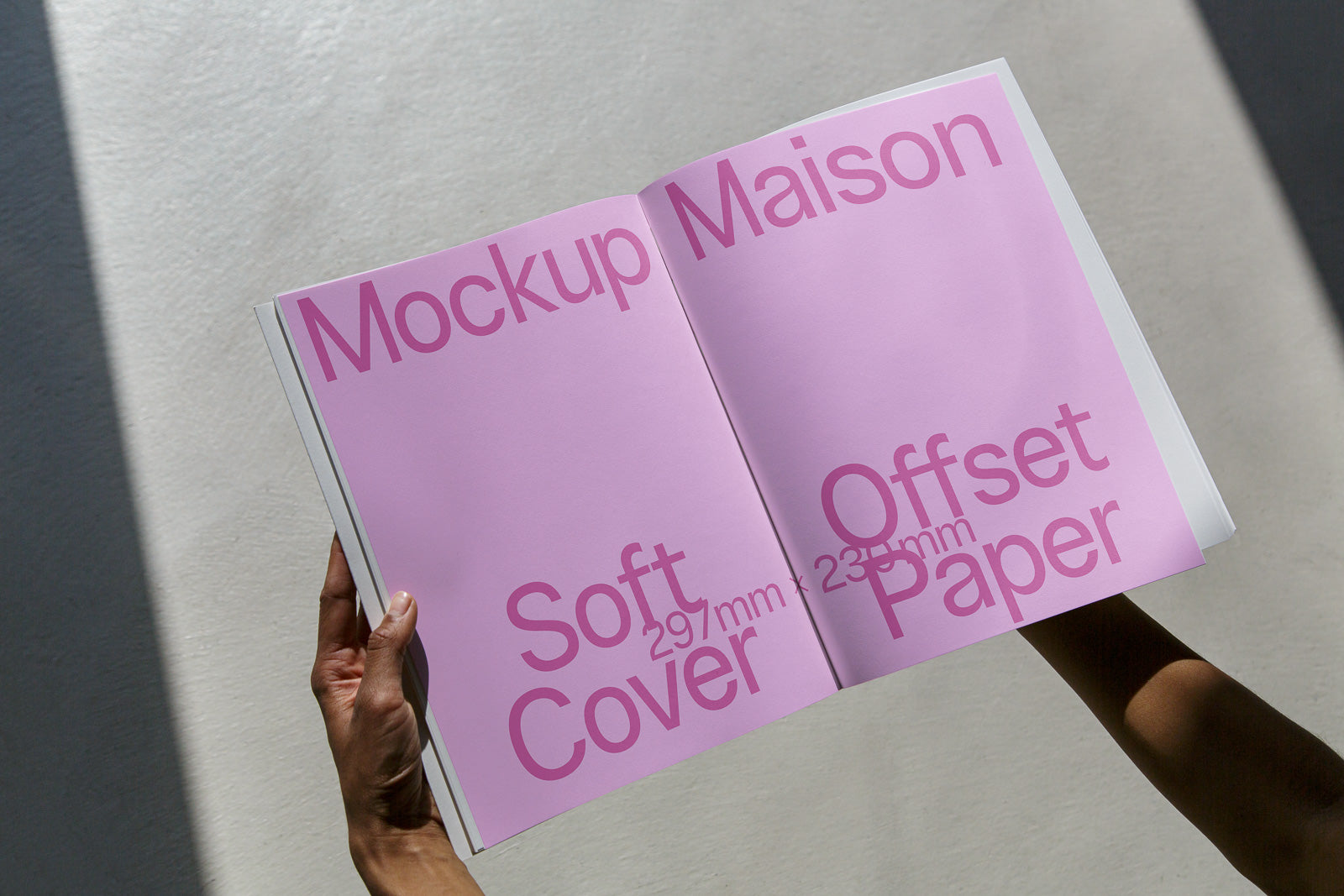 PSD mockup of an open magazine held by hands against a light-colored background, featuring pink pages with the words "Mockup Maison," "Offset Paper," and "Soft Cover."