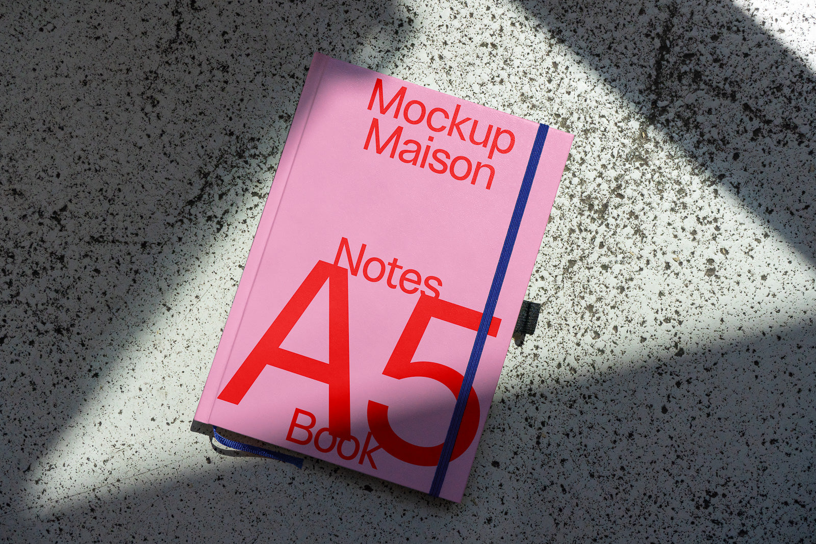 PSD mockup of a pink A5 notebook with red text on the cover, lying on a textured surface, partially shadowed.