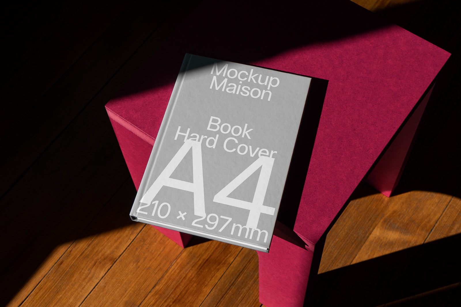 PSD mockup of an A4 hard cover book placed on a magenta stool with wood flooring background.