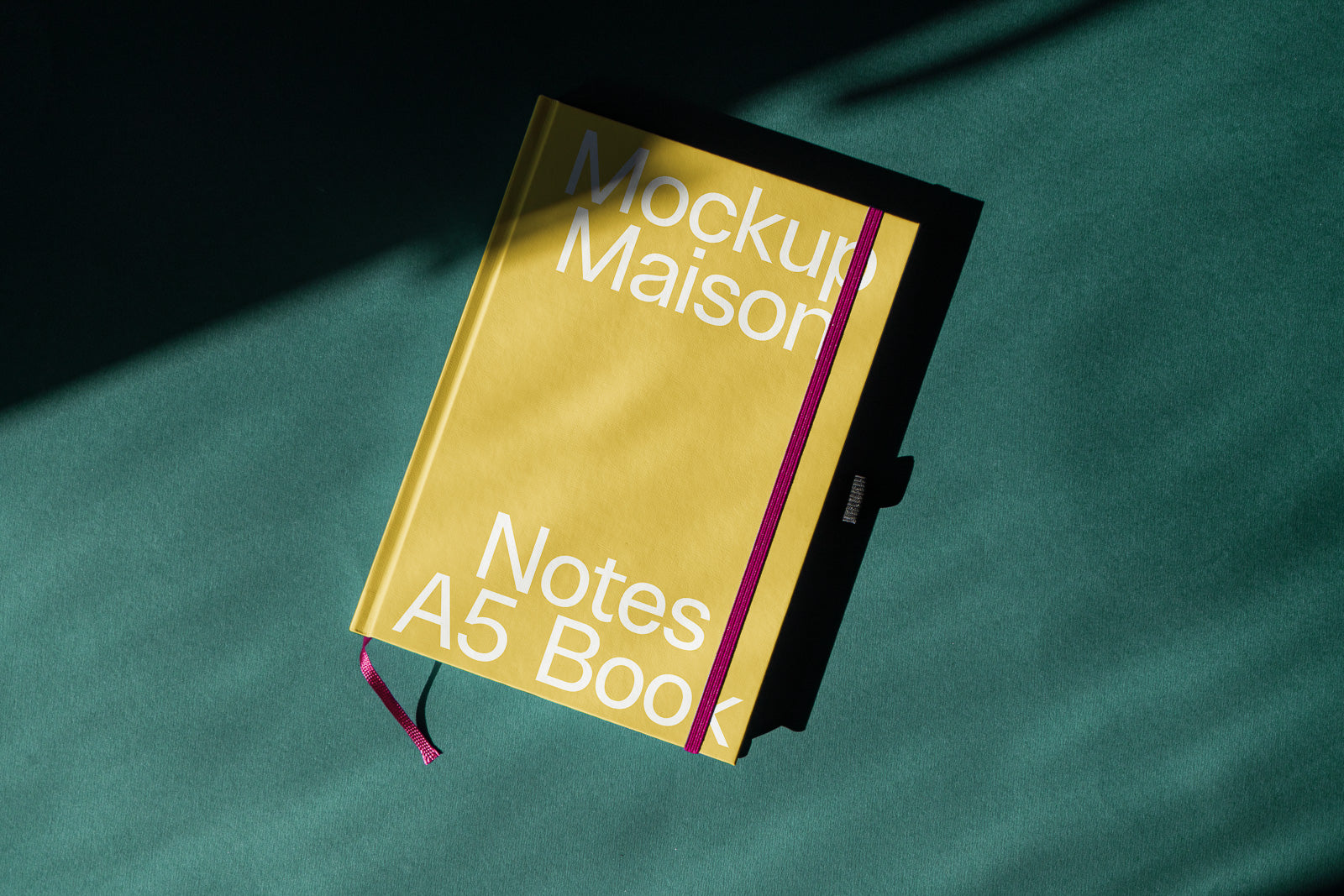 PSD mockup of a yellow A5 notebook with "Mockup Maison Notes A5 Book" text on the cover, featuring a pink elastic band and green background with shadows.