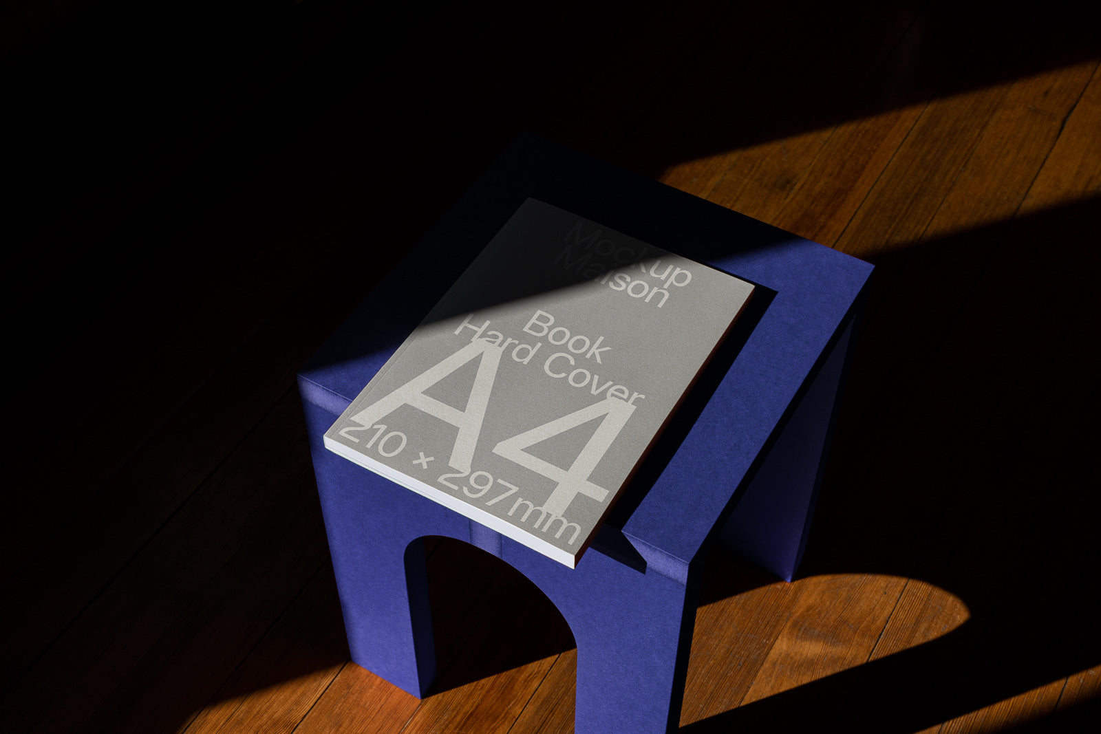 PSD mockup of an A4 hardcover book on a blue stool, with shadows and sunlight highlighting the cover's text and details.