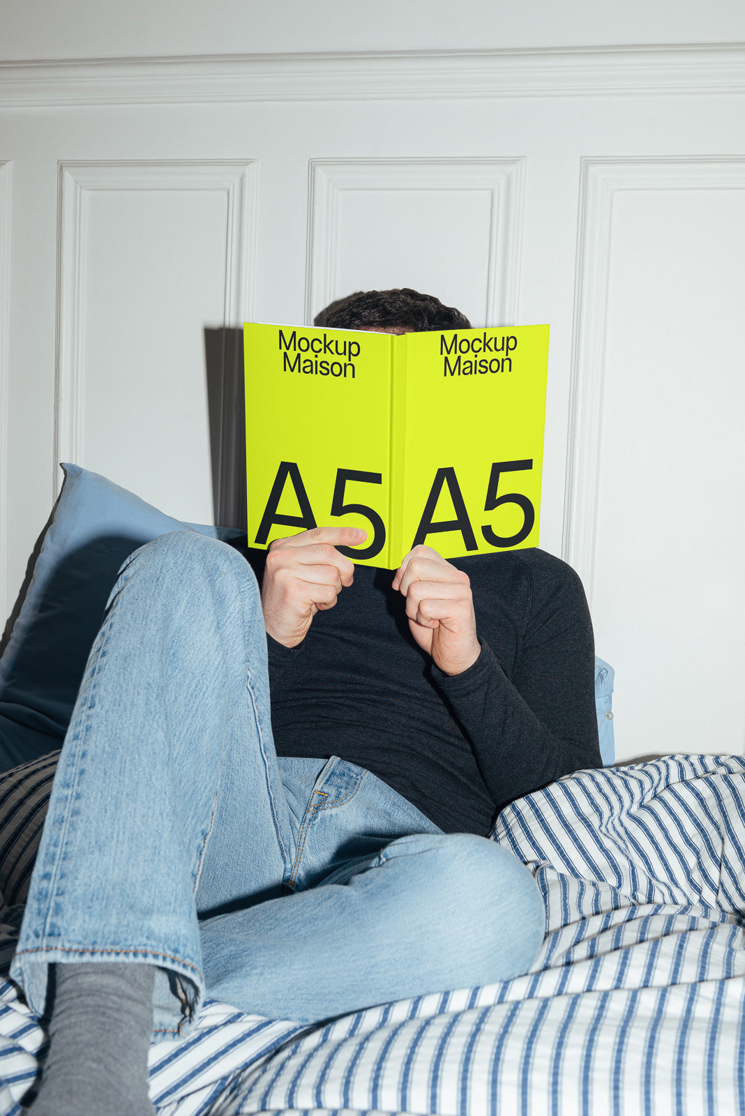 PSD mockup of a person relaxing on a bed, holding an open neon yellow book labeled "Mockup Maison A5," with a striped bedding in the background.