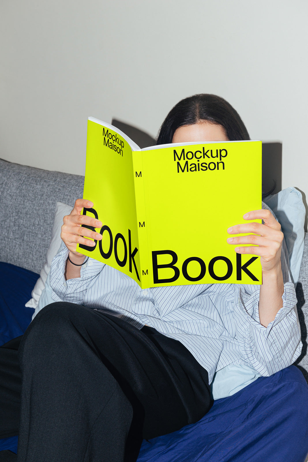PSD mockup of a person holding a bright yellow book titled "Mockup Maison" while sitting on a couch.