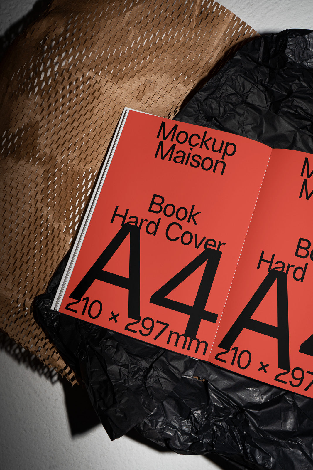 PSD mockup of an open A4 hardcover book with “Mockup Maison” text on a red page, placed on textured brown and black wrapping paper.
