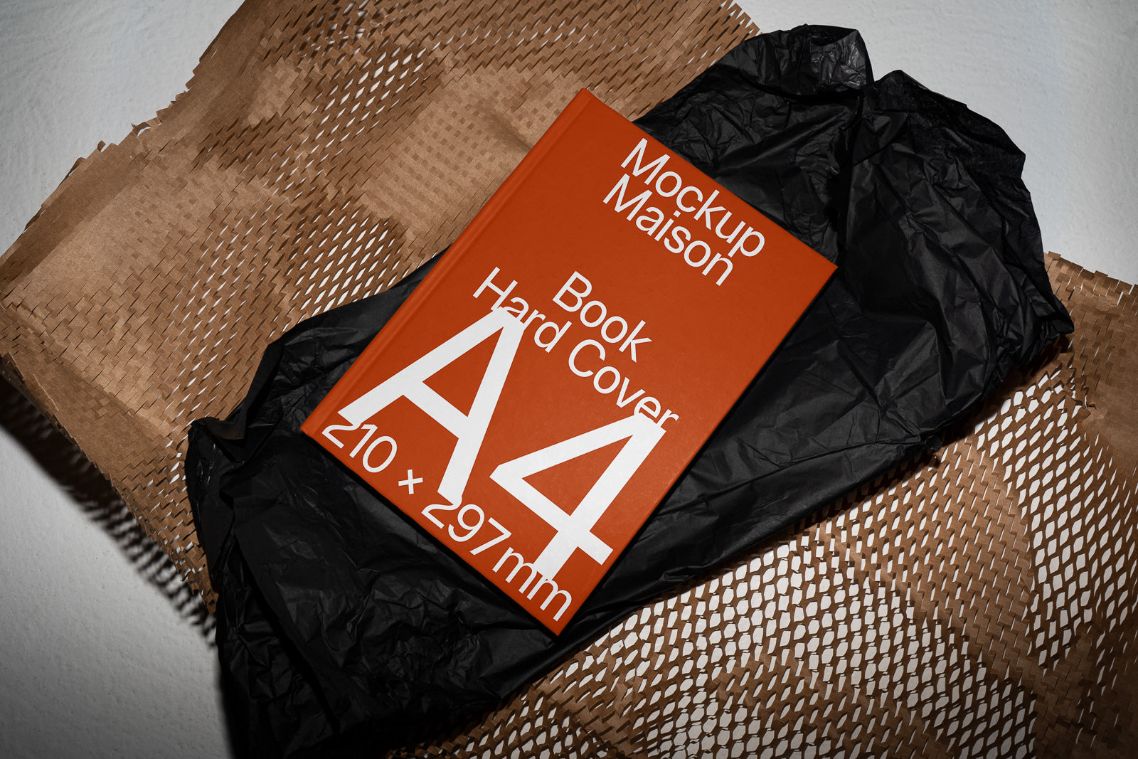 PSD mockup of a hard cover A4 book with "Mockup Maison" text on the cover, placed on textured brown and black wrapping paper.