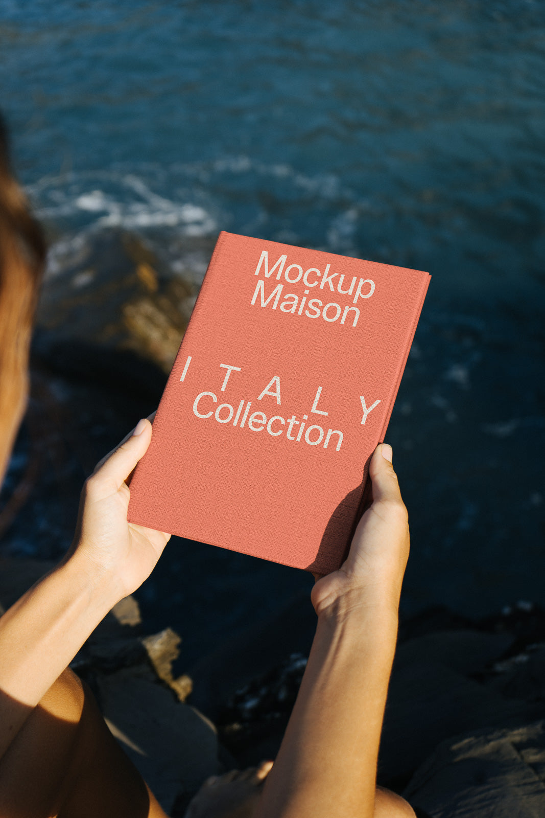 PSD mockup of a person holding a red book titled "Mockup Maison Italy Collection" near a rocky shore with water in the background.