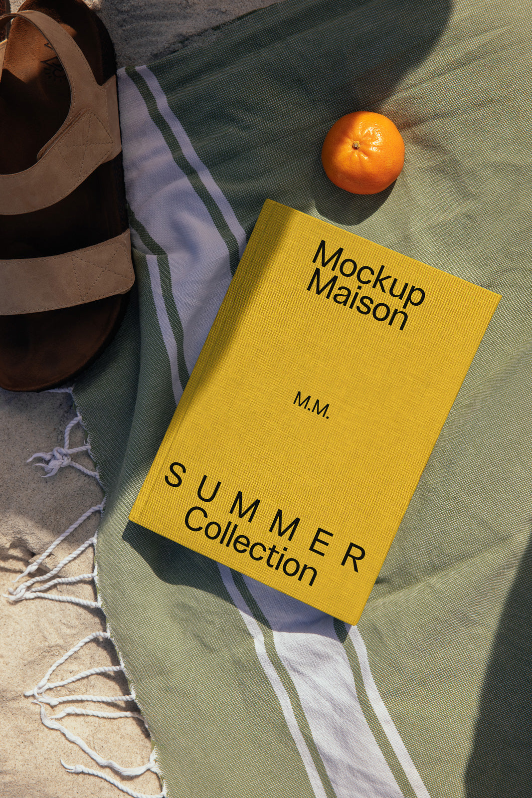 PSD mockup of a yellow book titled "Mockup Maison Summer Collection" on a striped cloth near a sandal and an orange.