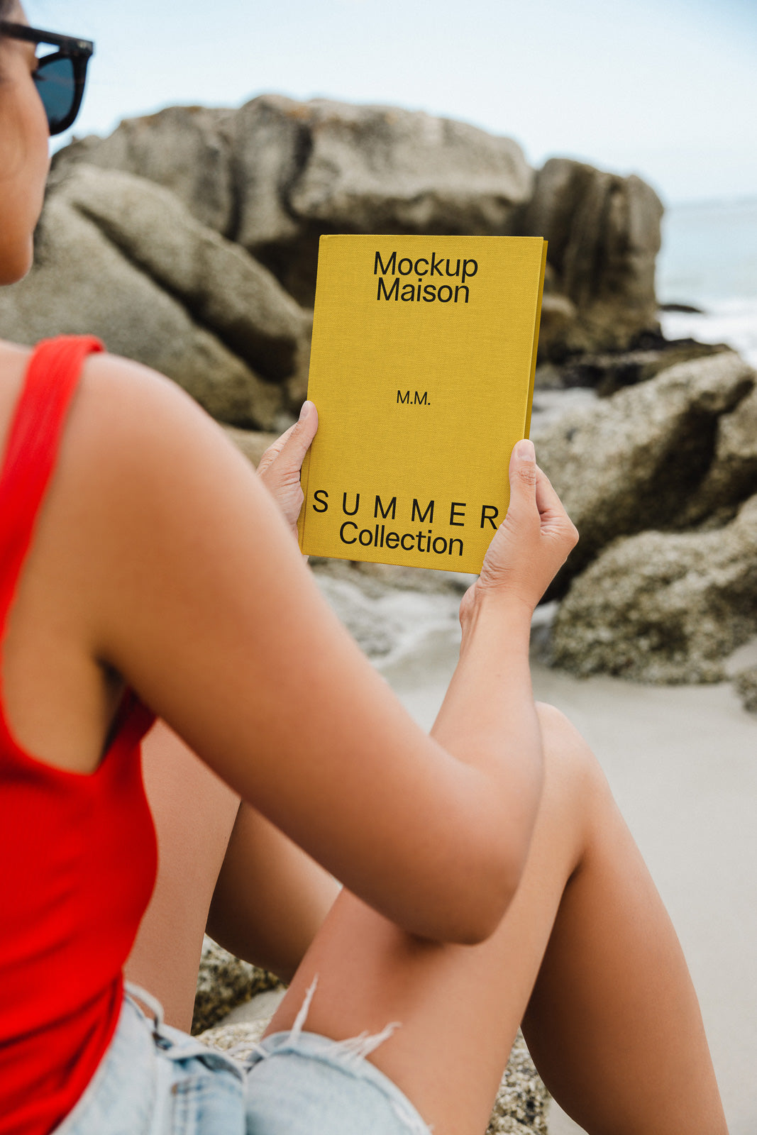 PSD mockup of a person on a beach holding a yellow book titled "Summer Collection" by "Mockup Maison," with ocean and rocks in the background.