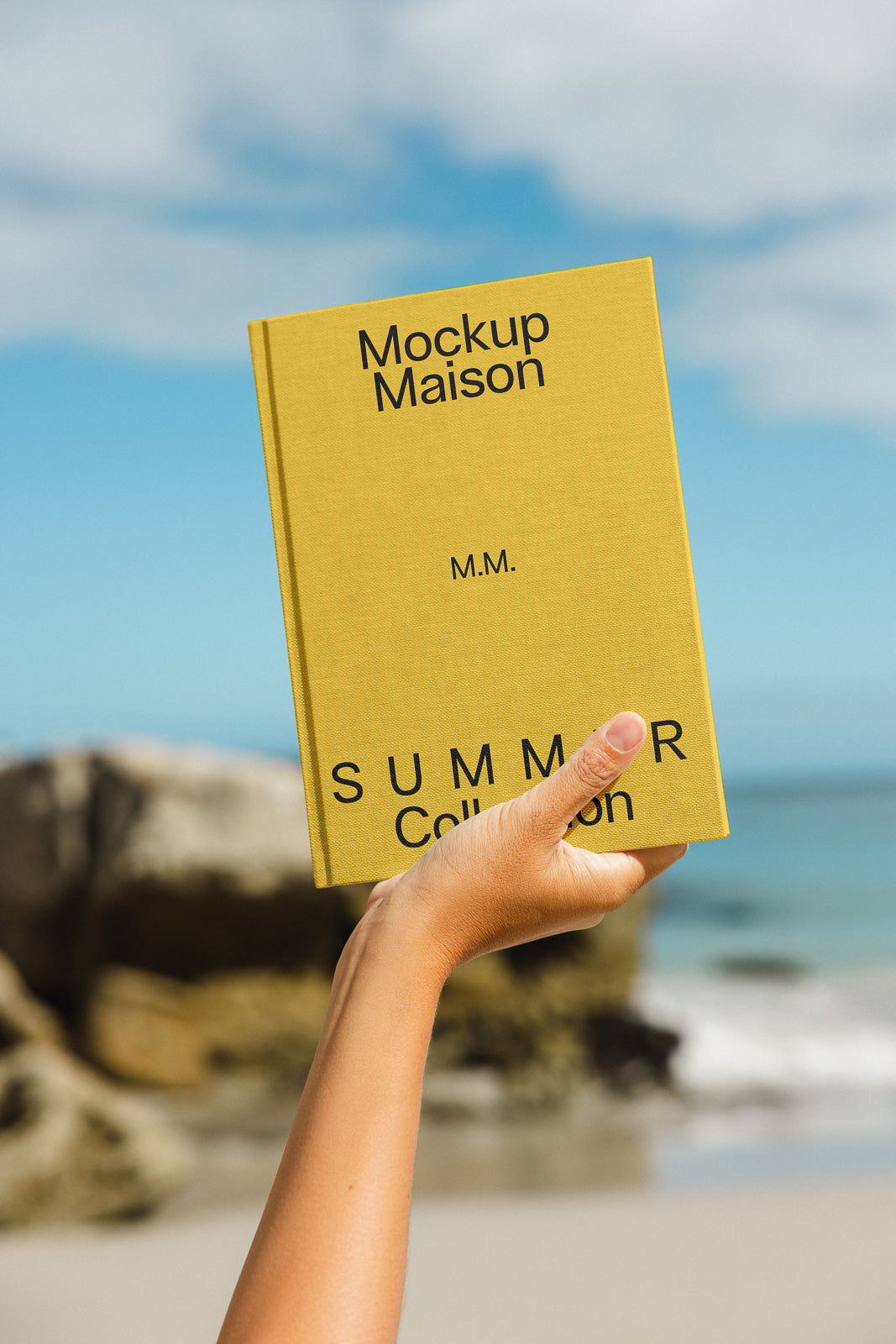 PSD mockup of a person holding a yellow book labeled "Mockup Maison, Summer Collection" against a beach background with a clear blue sky.