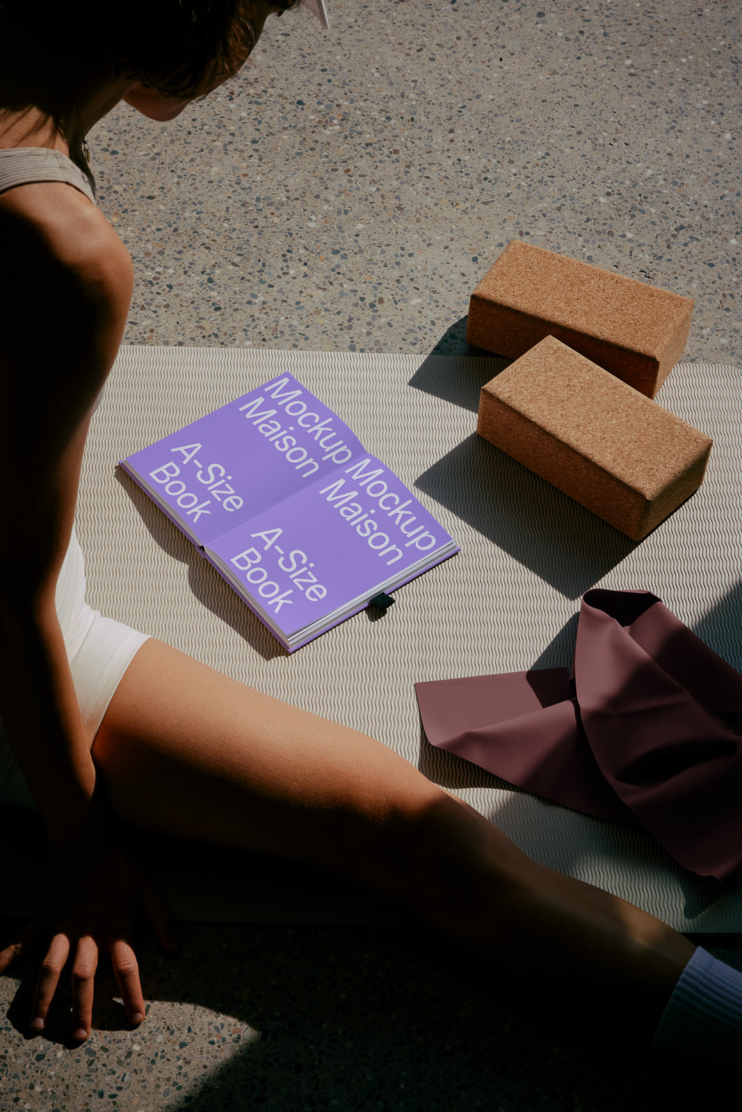PSD mockup of a purple A-size book open on a striped mat, accompanied by two cork yoga blocks, a dark mauve cloth, and a person sitting nearby on a sunny floor.