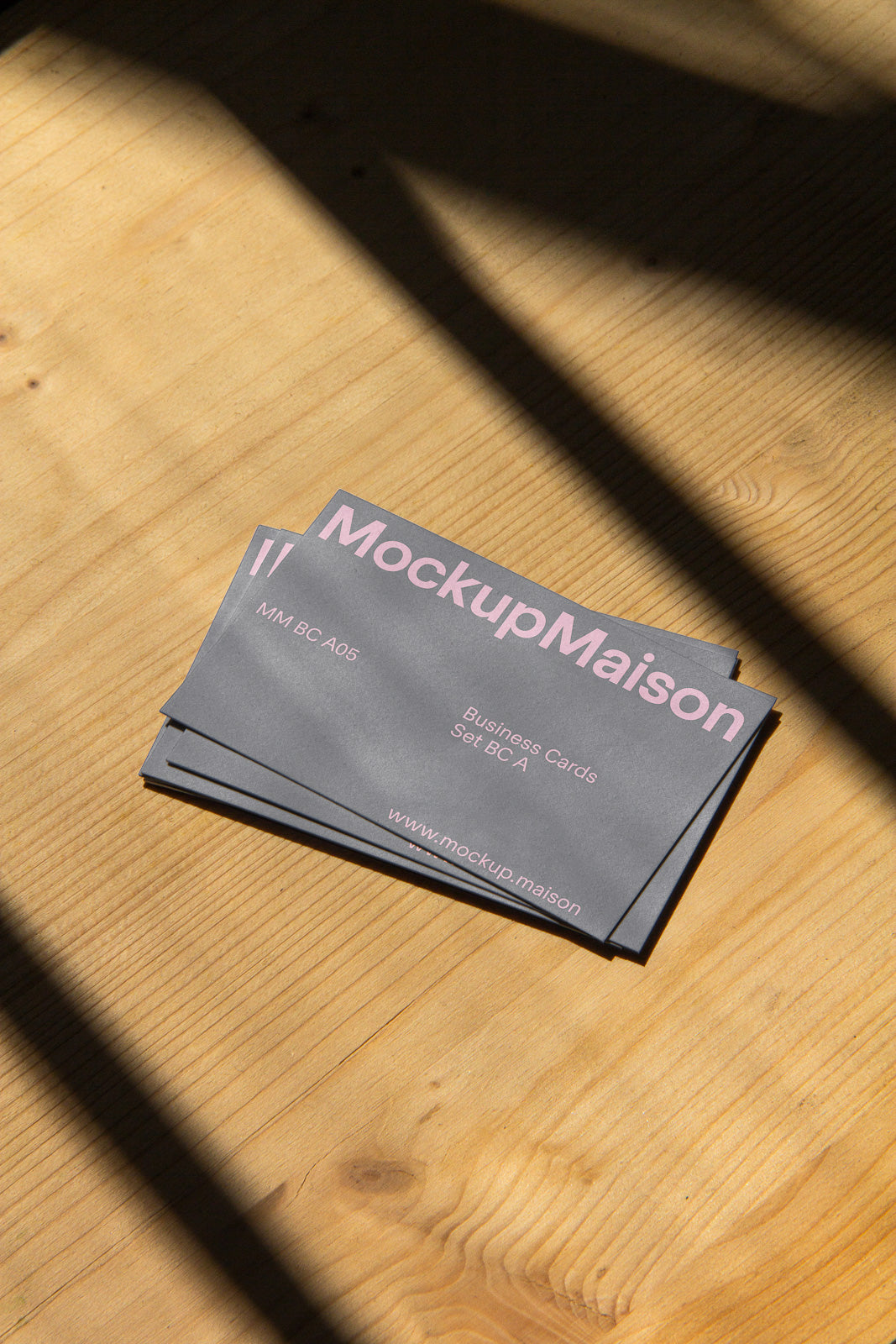 PSD mockup of stacked business cards with the text "MockupMaison" in pink on a wooden table, casting shadows across the surface.