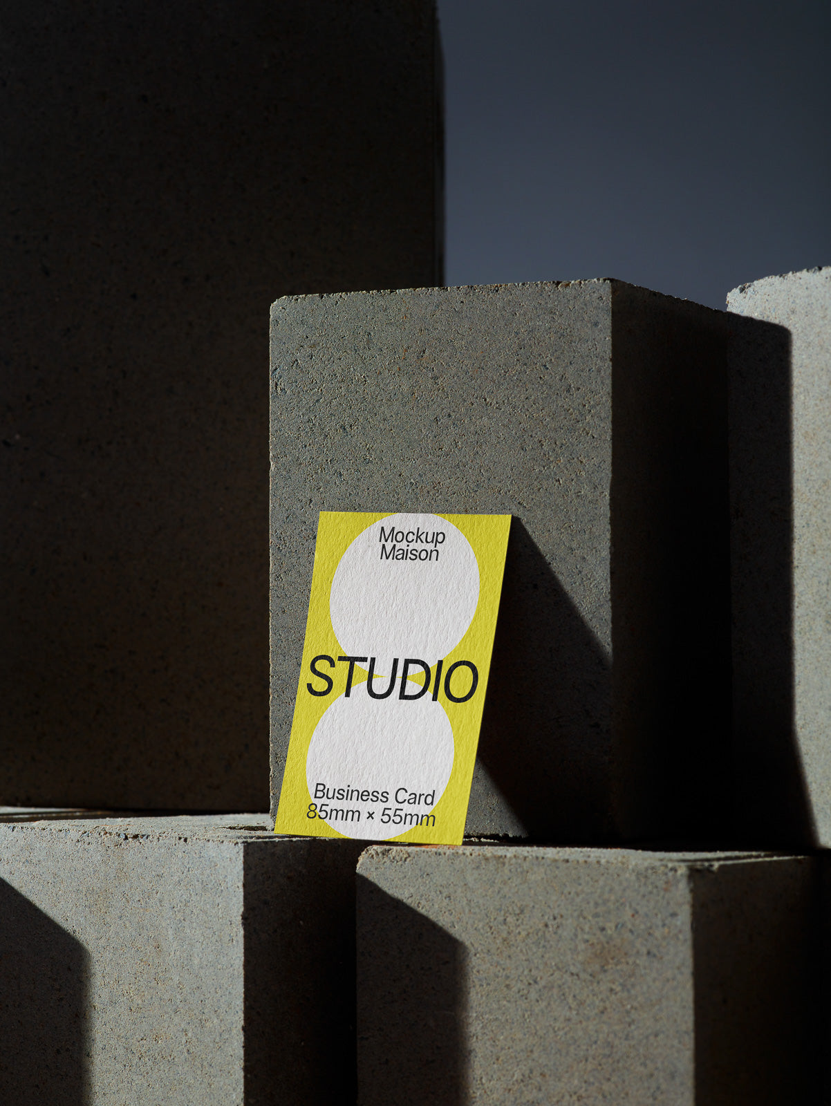 PSD mockup of a yellow-edge business card with bold "STUDIO" text, leaning against textured concrete blocks in dramatic lighting.