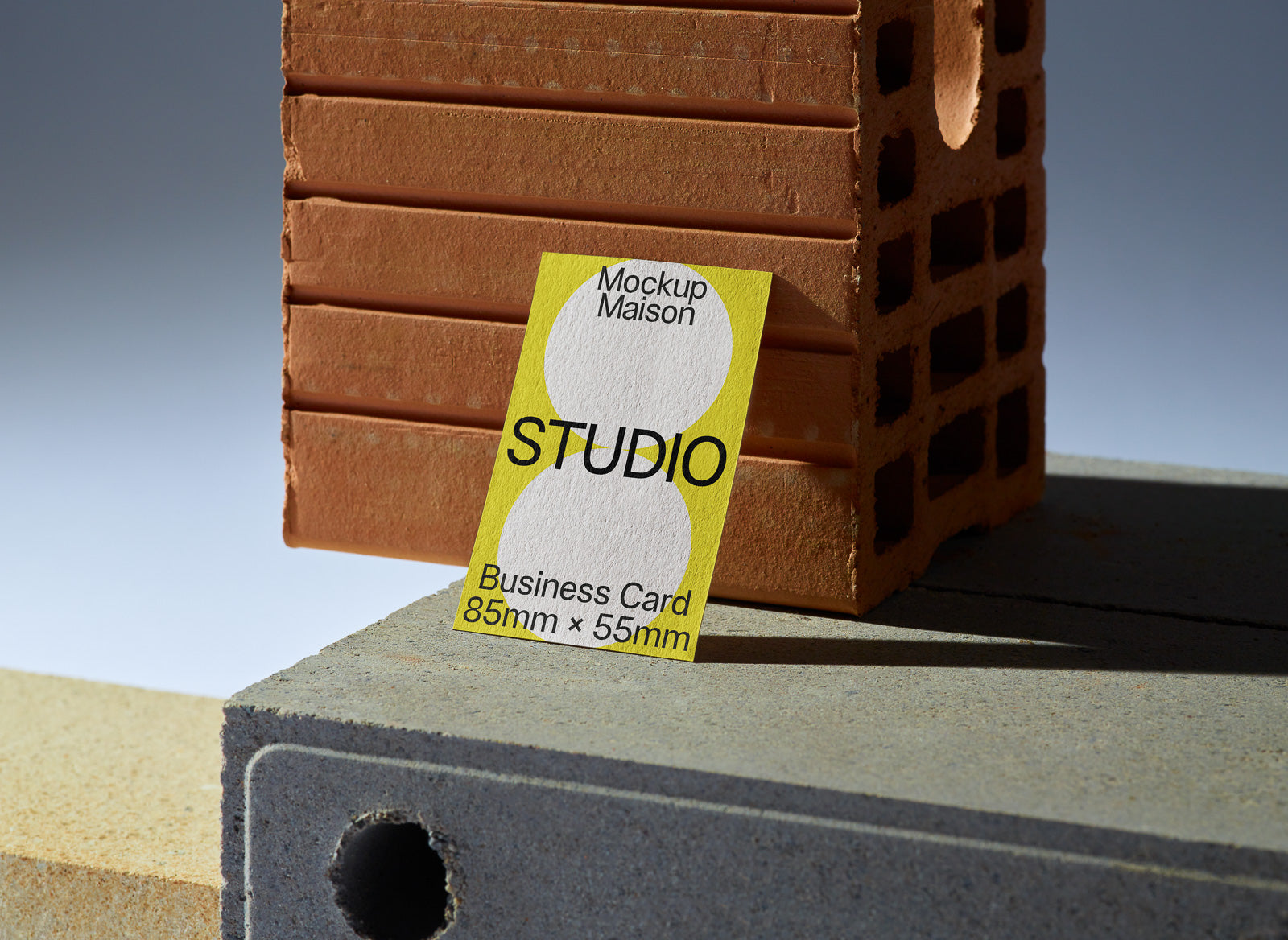 PSD mockup of a business card with "Mockup Maison," "STUDIO," and dimensions 85mm x 55mm in bold text, leaning against a textured brick surface.