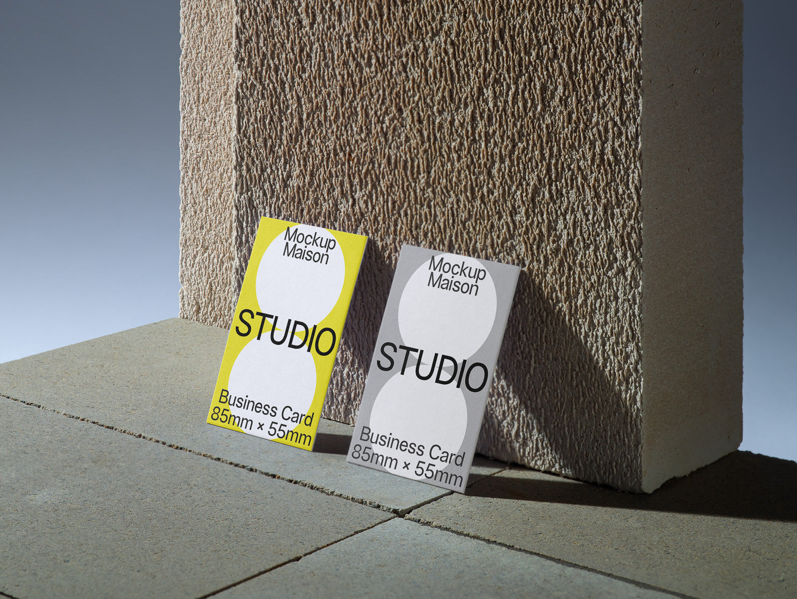 PSD mockup of two business cards, one yellow and one white, standing against a textured concrete block. Each card is labeled "Mockup Maison Studio" with dimensions 85mm x 55mm.