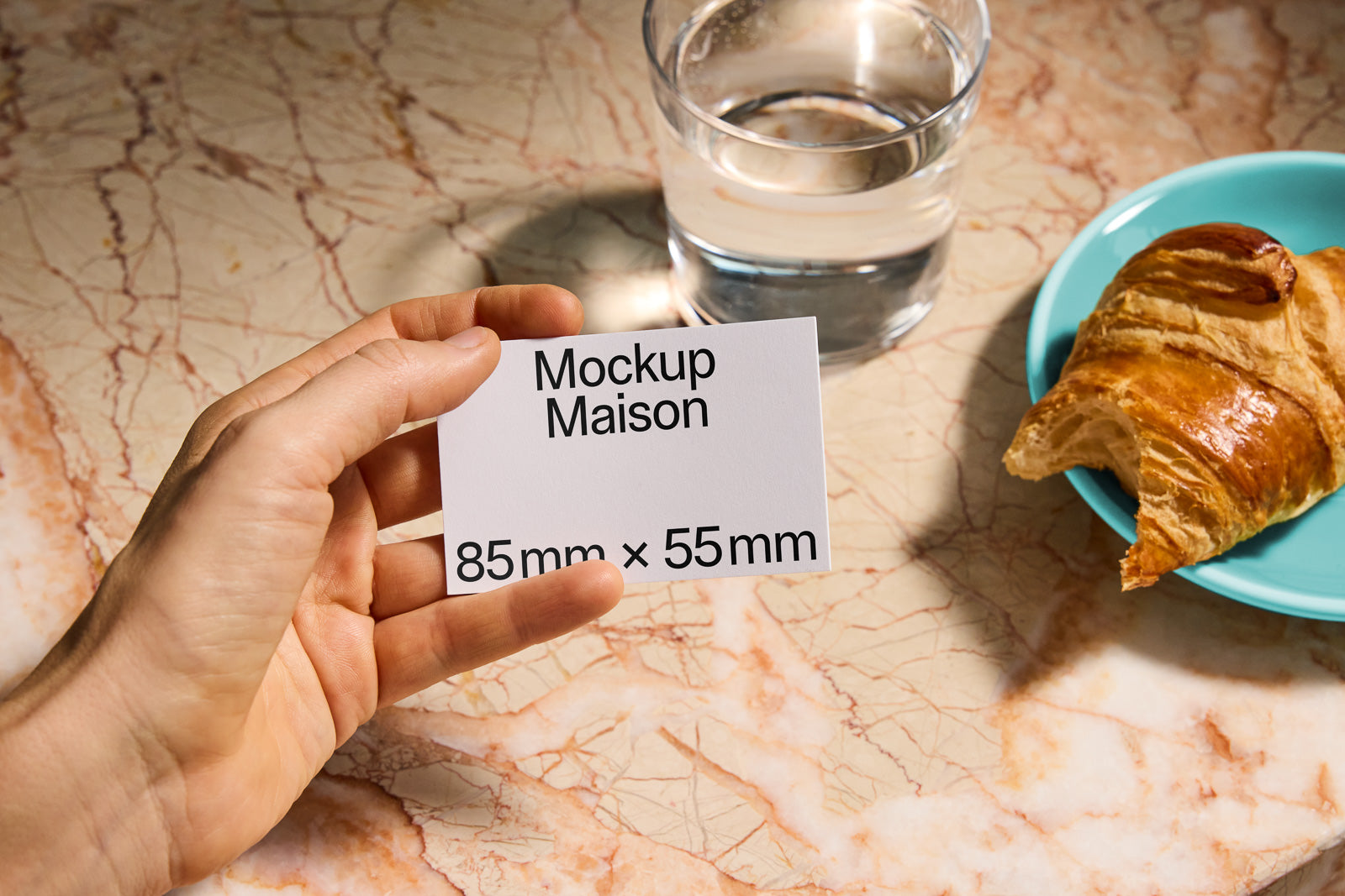 PSD mockup of a hand holding a business card labeled "Mockup Maison 85mm x 55mm," placed on a marble surface near a glass of water and a croissant on a blue plate.