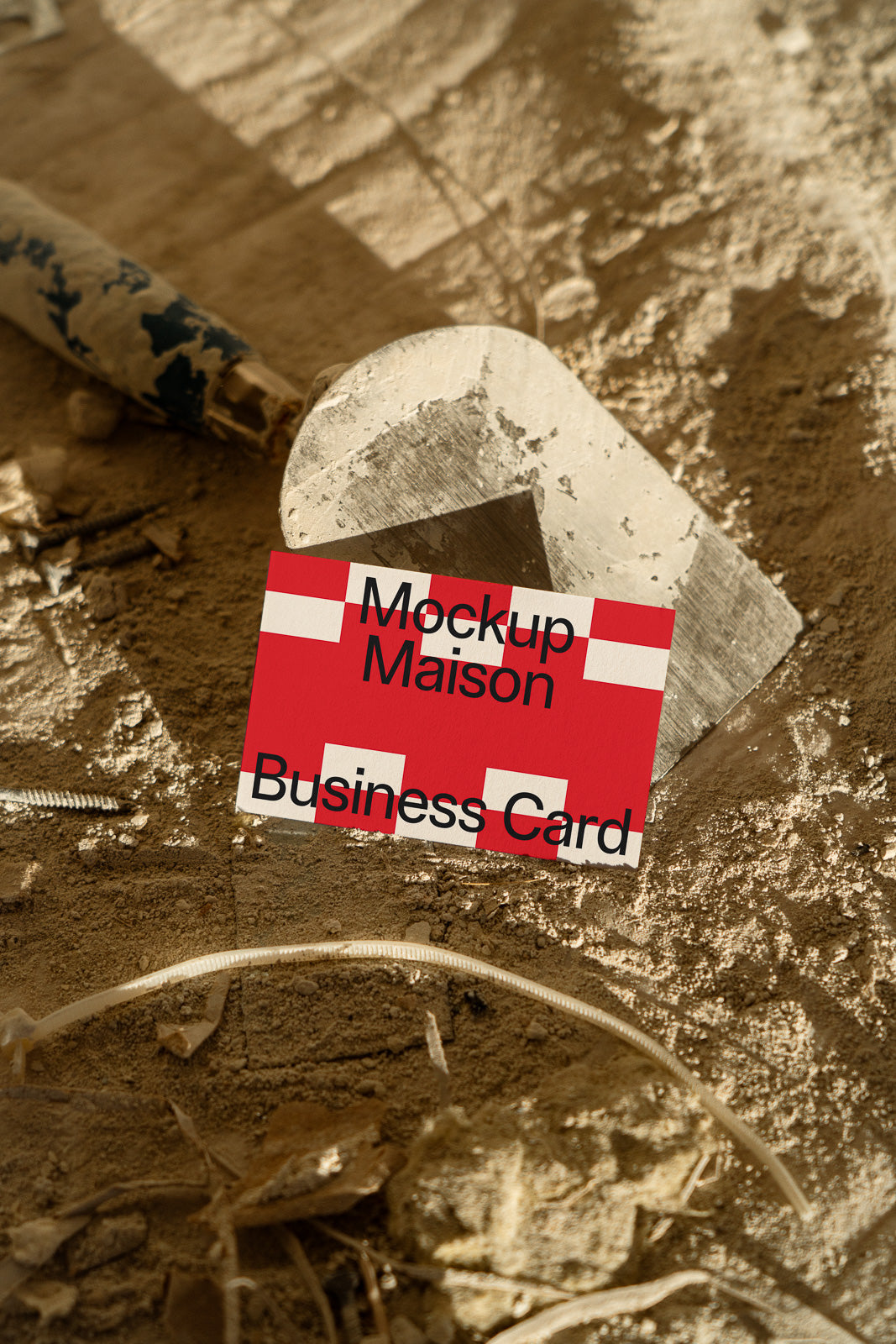 PSD mockup of a red business card with white crosses and text, placed on a rough, dusty surface with construction tools in the background.