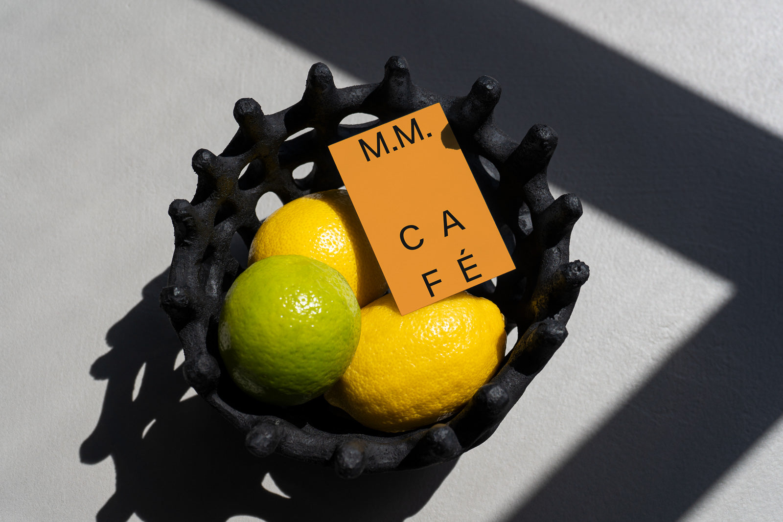 PSD mockup of a business card labeled "M.M. Café" resting on lemons and a lime in a textured black bowl, with a shadow cast across a gray surface.