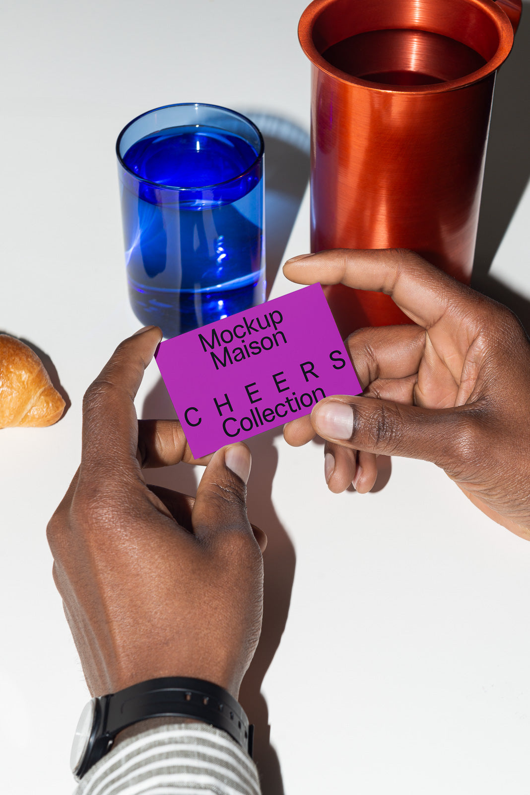 PSD mockup of hands holding a purple card labeled "Mockup Maison Cheers Collection" near a blue glass, red cup, and a croissant on a white table.