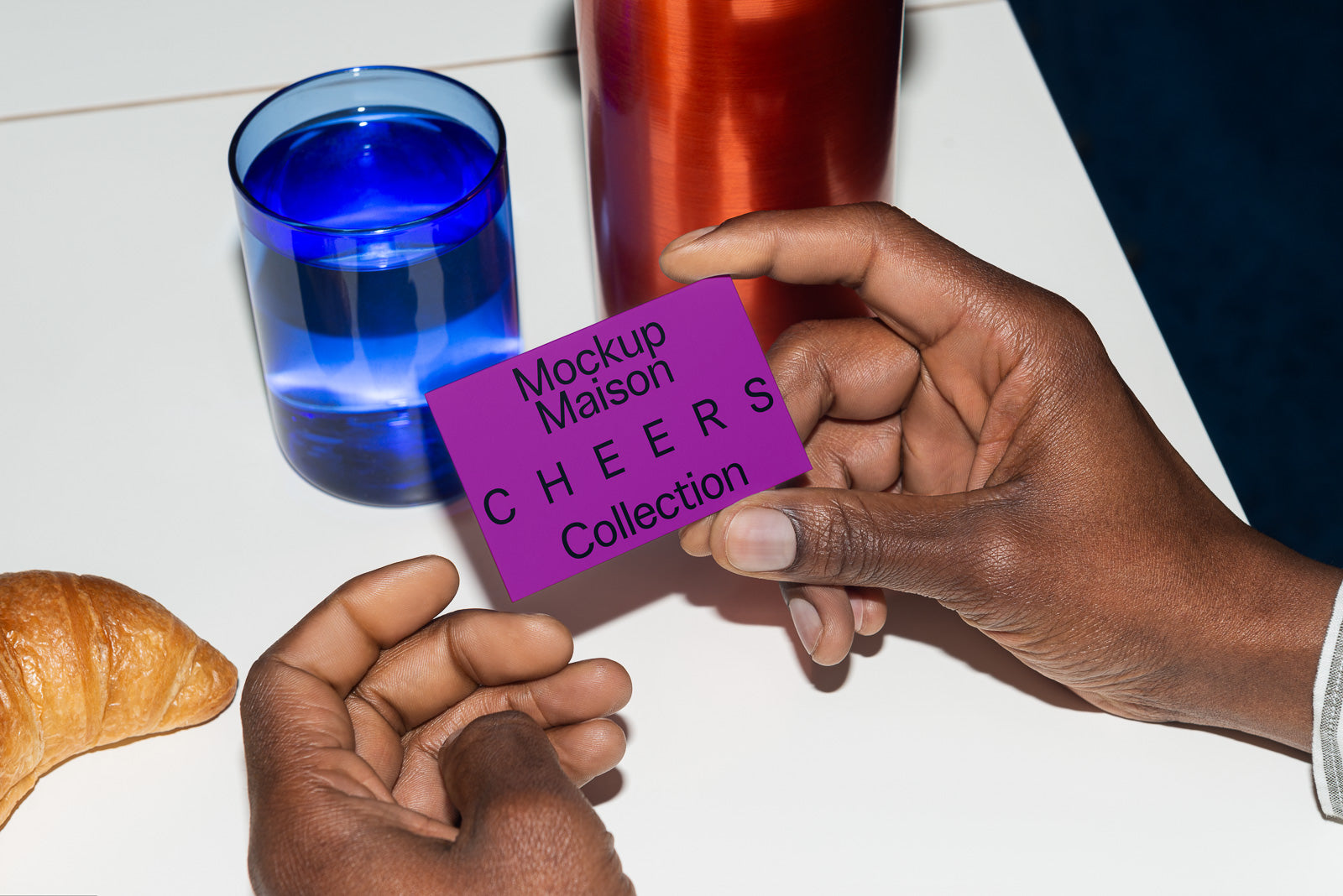 PSD mockup of hands holding a purple card with "Mockup Maison CHEERS Collection" near a blue glass, red bottle, and a croissant on a white surface.