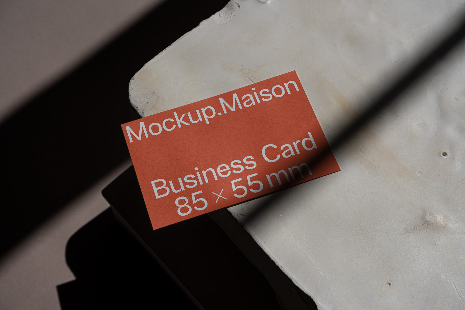 PSD mockup of a business card sized 85x55 mm on a textured surface with shadows, highlighting "Mockup.Maison" in white text on an orange background.