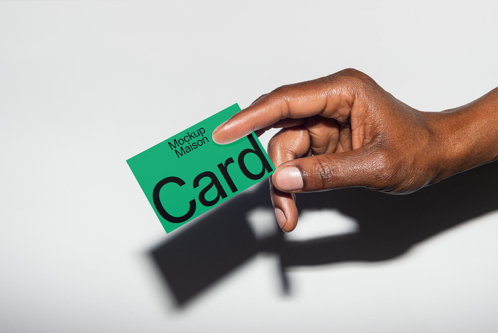 PSD mockup of a hand holding a green card with "Mockup Maison" text, placed against a light surface, casting a shadow below.