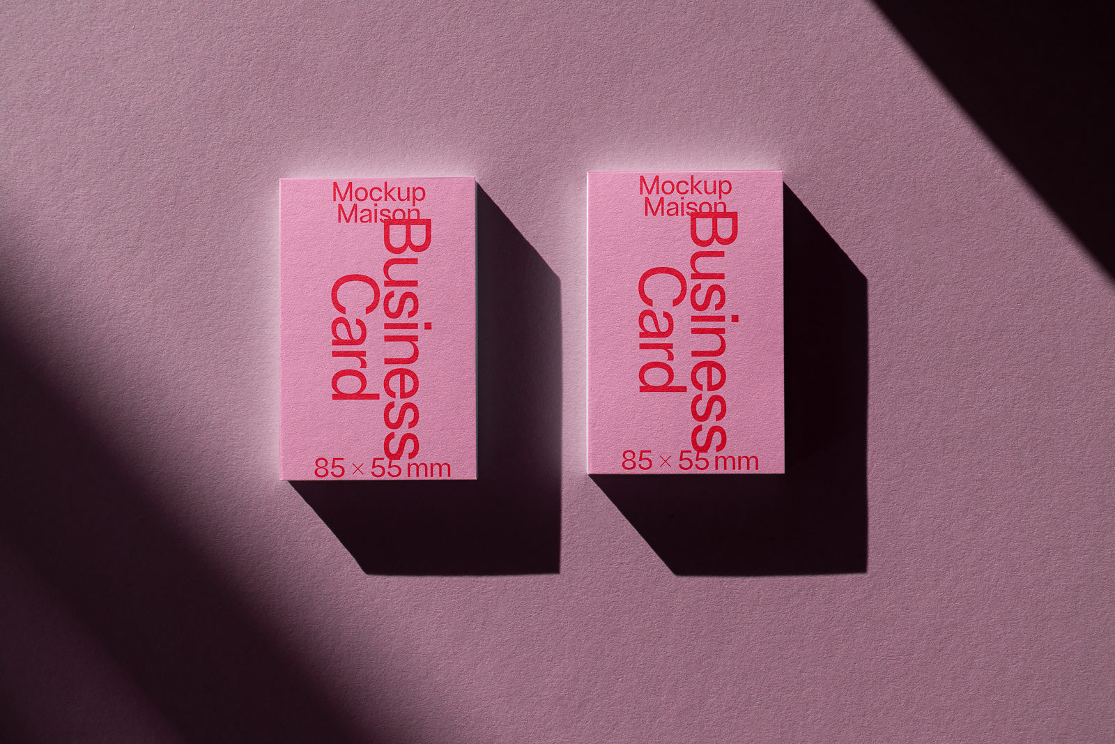 PSD mockup of two pink business cards with red text and dimensions 85×55mm on a textured pink surface, casting soft shadows.